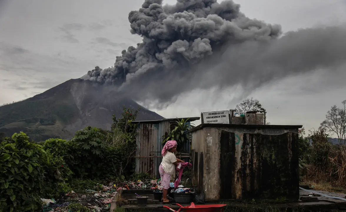 14 August 2020, Indonesia, Karo: A woman washes her clothes as Mount Sinabung Volcano spews thick ash and smoke into the sky following its recent eruption. Photo: Albert Ivan Damanik/dpa.