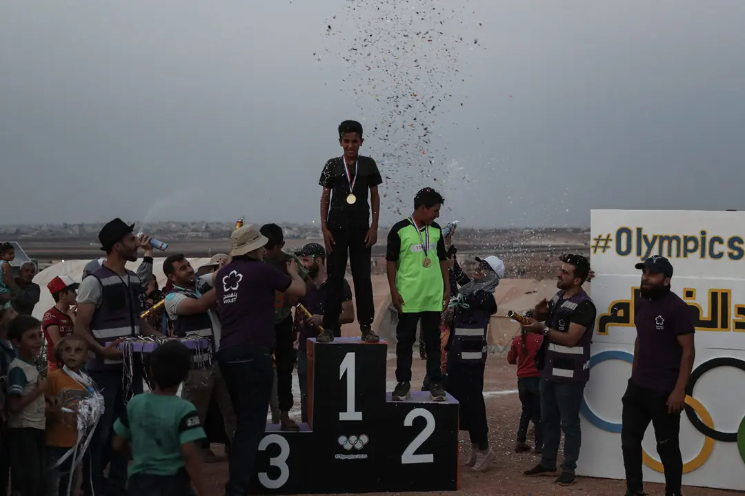 07 August 2021, Syria, Idlib City: Syrian children celebrate on the podium with symbolic medals distributed among the winners of a small-scale Olympic-style event held for internally displaced children by volunteers of the Violet relief organization. The sports event is organized in conjunction with the Tokyo 2020 Olympic Games to draw attention to the conditions inside the refugee camps of those who were forced into internal displacement as a result of the Syrian civil conflict. Photo: Anas Alkharboutli/dpa