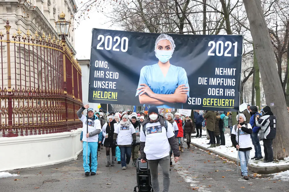 11 December 2021, Austria, Vienna: Demonstrators hold a banner showing a health worker between the phrases "2020 OMG you are our heroes..2021 take the vaccine or you are fired" as they take part in a protest against the Corona measures and the planned mandatory vaccination. Photo: Florian Wieser/APA/dpa,