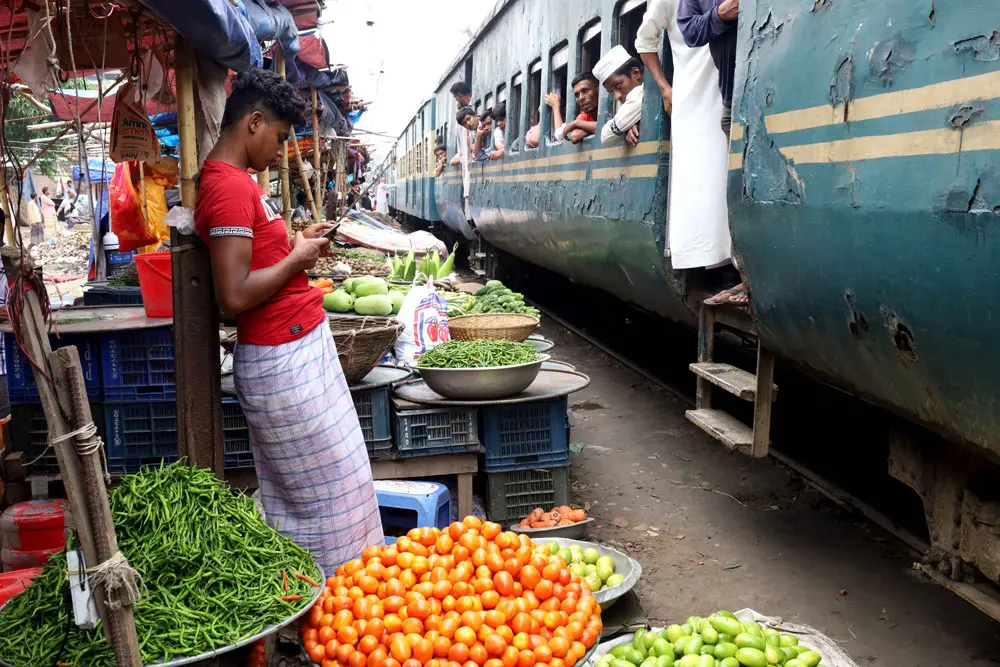 16 June 2022, Bangladesh, Dhaka: A train passes through the Jurain Railgate market just inches from people selling fruit and vegetables. Two trains pass through this market at a speed of about 30 kilometres per hour. Photo: Syed Mahabubul Kader/ZUMA Press Wire/dpa.