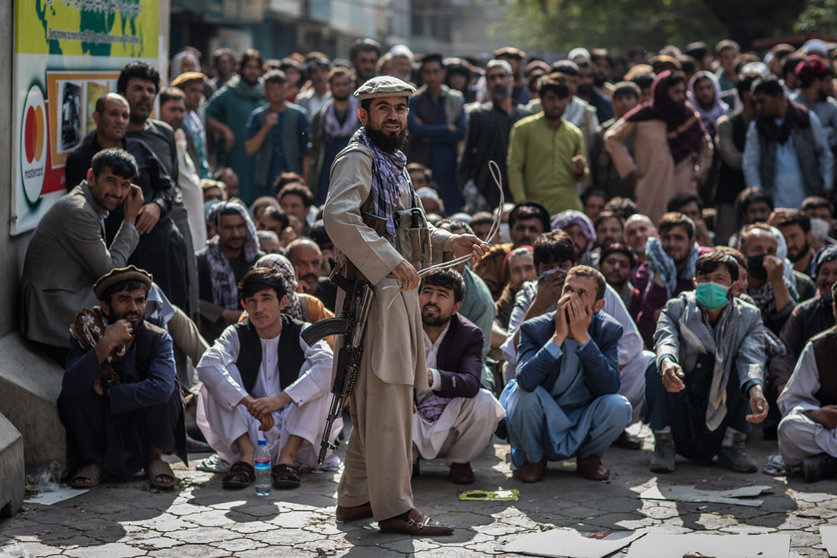 A Taliban security guard plays with a whip while standing in front of Afghan men waiting outside a bank to withdraw money. Photo: Oliver Weiken/dpa.