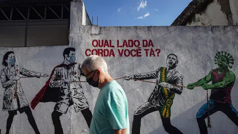 A man wears a face mask walks past by a mural depicting Brazilian President Jair Bolsonaro and a figure representing the coronavirus in a tug of war against health workers. Photo: Cris Faga/ZUMA Wire/dpa