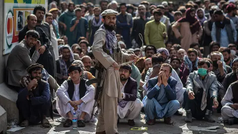 A Taliban security guard plays with a whip while standing in front of Afghan men waiting outside a bank to withdraw money. Photo: Oliver Weiken/dpa.